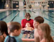 A swim instructor assists a child with a back float in an indoor pool. Text Overlay: Private Swimming Lessons, all ages