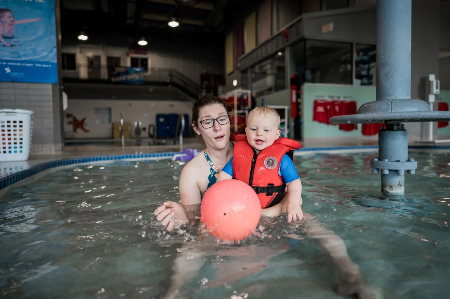 A woman holds a small child in an indoor pool. Text overlay: Drop in Schedule